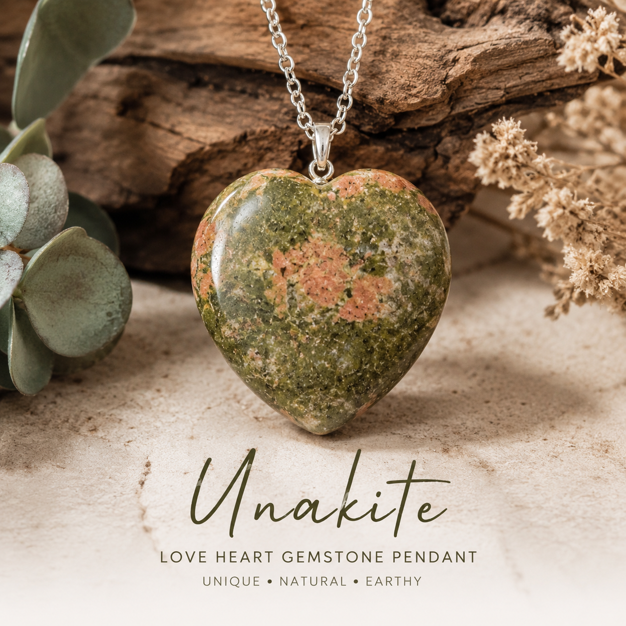 Heart-shaped unakite gemstone pendant necklace on a silver chain, styled with dried flowers and green leaves on a neutral surface, with text overlay reading 'Unakite LOVE HEART GEMSTONE PENDANT'.