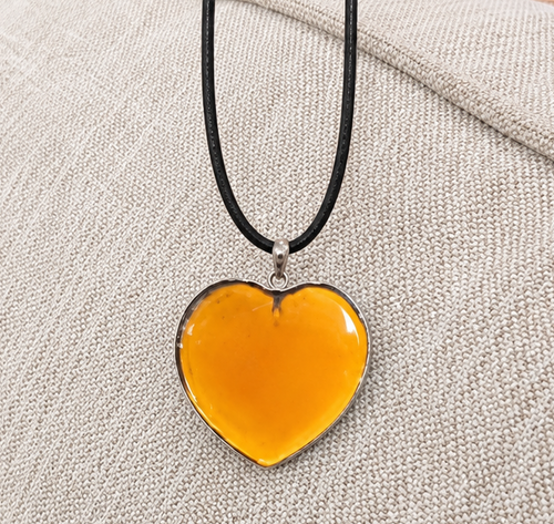 A heart-shaped amber glass pendant necklace with a silver metal border and black cord, resting on a textured beige fabric surface.